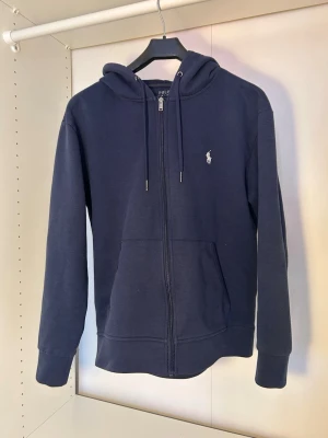 Mörkblå zip hoodie från Polo Ralph Lauren - Mörkblå hoodie med dragkedja från Polo Ralph Lauren. Klassisk design med huva och dragsko, två fickor framtill och den ikoniska broderade loggan på bröstet. Tillverkad i mjukt bomullsmaterial som är skönt att bära.