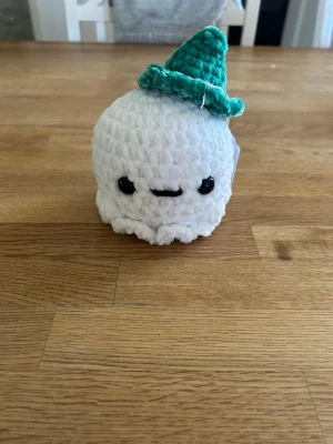 Virkad spöke med hatt - Söt handvirkad spöke i mjuk vit plysch med svarta säkerhetsögon. Spöket har en grön virkad hatt på huvudet och är perfekt som dekoration eller present. Liten och smidig att hålla i handen.