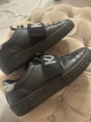 Svarta y2k  sneakers - Snygga svarta sneakers från Karl Lagerfeld med bred kardborreband dekorerat med glittrande stenar och texten KARL. Skorna har svarta snören, låg profil och silverfärgade detaljer vid hälen. Skosnörena är lite slitna av katdborrebandet. Nypris ligger kring 3500kr