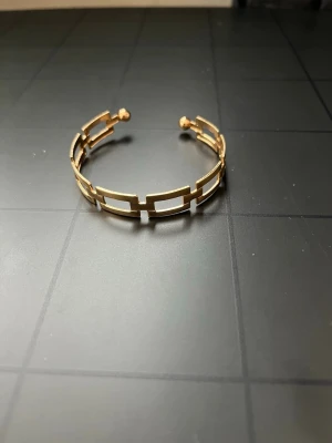 Guldigt öppet armband i metall - Snyggt armband i guldfärgad metall med öppen design och rektangulära länkar. Armbandet har två kulor i ändarna och en modern, stilren look som passar perfekt till din outfit.