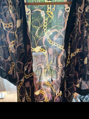 Svart kimono morgonrock med kedjemönster - Snygg svart morgonrock i kimonomodell med transparent tyg och mönster av guldfärgade kedjor och detaljer i rosa och beige. Långa, vida ärmar och knytband i midjan. Perfekt för en chill morgon eller kväll.