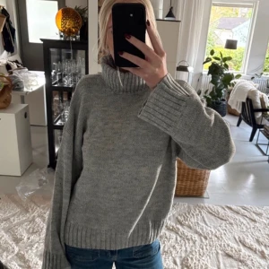 Grå polotröja i stickat - Mysig grå stickad polotröja med breda ärmar och ribbade muddar vid ärmslut och nederkant. Tröjan har en hög polokrage och en avslappnad passform, perfekt för kyliga dagar. Passar till jeans eller kjol för en enkel och snygg look.
