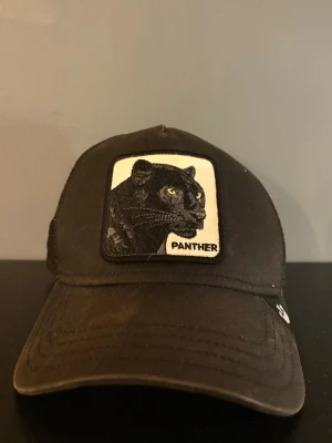 Svart keps med panterpatch - Svart keps med böjd skärm och mesh baktill för extra ventilation. Framtill sitter en broderad patch med en svart panter och texten 'PANTHER'. Justerbar storlek med plastspänne bak. 