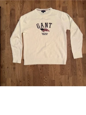 Vit stickad tröja från GANT - Vit stickad tröja från GANT med broderad logga och amerikansk flagga på bröstet. Tröjan har rund hals, långa ärmar och ribbstickade muddar vid ärmslut och nederkant. Perfekt för en stilren och klassisk look.