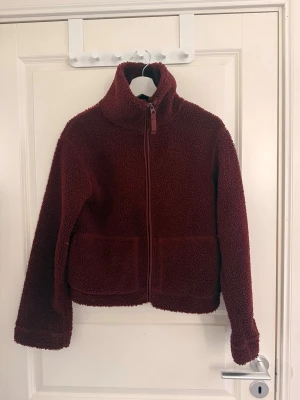 Vinröd teddyjacka från H&M - Mysig vinröd teddyjacka med hög krage och hel dragkedja framtill. Jackan har två stora fickor fram och är gjord i fluffigt teddy-material som håller dig varm. 