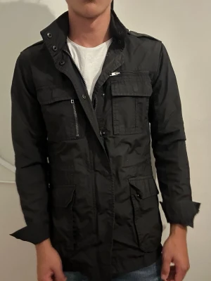 Svart field jacket med fickor - Stilren svart skepparkavaj med hög krage och flera praktiska fickor framtill, både med dragkedja och lock. Jackan har en rak passform och är tillverkad i ett lätt, tåligt material som passar perfekt till en modern look.