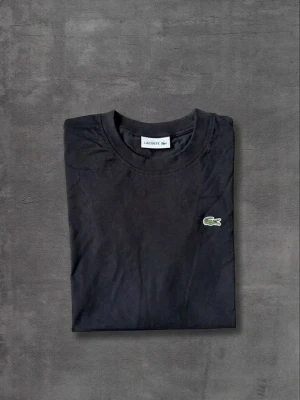 Svart t-shirt från Lacoste - Klassisk svart t-shirt från Lacoste med rund hals och den ikoniska gröna krokodilloggan broderad på bröstet. Tillverkad i mjuk bomull och har en normal passform. Perfekt basplagg för en clean och stilren look.