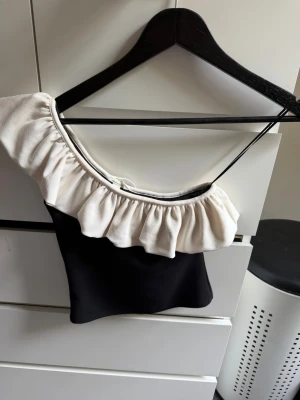 Svart och vit oneshoulder topp med volang - Trendig svart och vit oneshoulder topp med bred volang längs axeln. Toppen är i ett lite tjockare material. Skriv privat för fler bilder eller frågor 💕