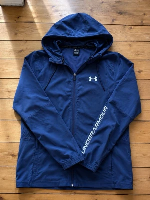 Blå vindjacka från Under Armour - Snygg marinblå vindjacka från Under Armour med huva och dragkedja framtill. Jackan har logga på bröstet och texten UNDER ARMOUR på ena ärmen. Tillverkad i lätt polyester, perfekt för blåsiga dagar.
