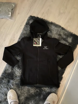 Svart vindjacka från Arc'teryx - Snygg svart vindjacka från Arc'teryx med huva och dragkedja framtill. Jackan har två sidofickor med dragkedja och är tillverkad i slitstarkt GORE-TEX-material. Perfekt för blåsiga dagar och outdoor-äventyr.