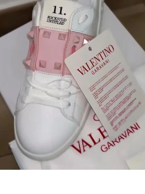 Valentino Rockstud Untitled sneakers - Snygga vita sneakers från Valentino Garavani med rosa band och ikoniska nitar över snörningen. Skorna har en låg silhuett och är tillverkade i läder med guldfärgad logga på innersulan. Perfekta för dig som vill sticka ut med exklusiv design.
