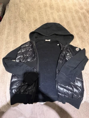 Svart pufferjacka med huva från Moncler - Snygg svart pufferjacka från Moncler med glansiga paneler framtill och baktill, samt mjuka ärmar och huva i sweatshirtmaterial. Jackan har dragkedja, ribbade muddar och Moncler-logga på ärmen. Perfekt för kalla vinterdagar.
