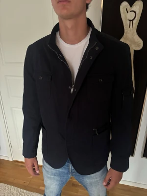 Svart jacka med dragkedja och fickor - Snygg svart jacka med dragkedja framtill och flera fickor med lock och dragkedja. Jackan har en klassisk krage och lång ärm, perfekt för en stilren look. Passar till jeans och t-shirt för en avslappnad stil.
