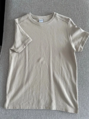 Beige ribbad t-shirt i bomull - Säljer en stilren beige t-shirt med ribbad struktur och rund hals. T-shirten har korta ärmar och är tillverkad i mjuk bomull, perfekt för en enkel och clean look.
