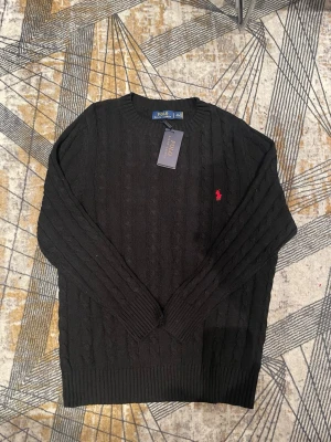 Svart kabelstickad tröja Polo Ralph Lauren - Kabelstickad svart tröja från Polo Ralph Lauren med rund halsringning och den klassiska röda logotypen broderad på bröstet. Tröjan har långa ärmar och ribbade muddar vid ärmslut och nederkant. Perfekt för en stilren och tidlös look.