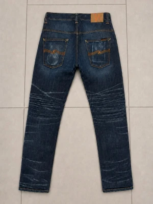 Nudie Jeans 29/34 - Säljer ett par mörkblå jeans med snygga slitningar och tvättade detaljer på både fram- och baksida. Jeansen har raka ben, klassiska fem fickor och kontrastsömmar. Bakfickorna har dekorativa sömmar i orange. Perfekta för en avslappnad stil.