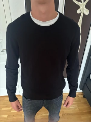 Svart basic tröja med rund hals - En stilren svart långärmad tröja med rund halsringning. Tröjan har en enkel och clean design utan synliga loggor eller mönster, perfekt att matcha med olika outfits. Tillverkad i ett mjukt material som sitter skönt på kroppen.