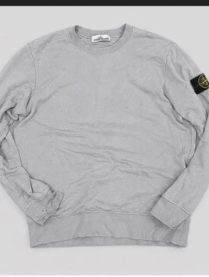 Grå sweatshirt från Stone Island - Säljer en stilren grå sweatshirt från Stone Island med klassisk rund hals och ribbade muddar. Tröjan har den ikoniska svarta patchen med gul och grön logga på vänster ärm. Perfekt för dig som gillar streetwear och enkel design.