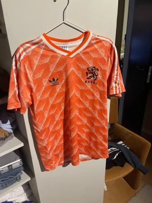 Orange Holland fotbollströja Adidas - Säljer en ikonisk orange Holland fotbollströja från Adidas med vitt geometriskt mönster, vita ränder på axlarna och KNVB-logga på bröstet. Tröjan har V-ringning med vit och orange kant samt korta ärmar. Perfekt för dig som älskar retro sportstil.