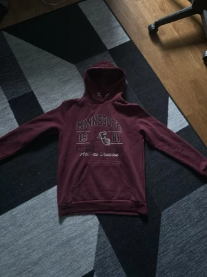 Vinröd hoodie med Minnesota-tryck - Snygg vinröd hoodie med tryck 'Minnesota 1981 North State Athletic Division' och en hjälm på bröstet. Klassisk känguruficka framtill och huva med dragsko. Vintage hoodie. Pris kan diskuteras 