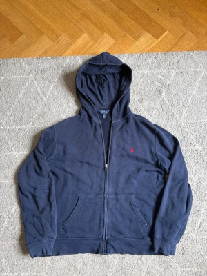 Mörkblå hoodie från Polo Ralph Lauren - Mörkblå hoodie med dragkedja från Polo Ralph Lauren. Klassisk design med huva, kängurufickor och röd broderad logga på bröstet. Tillverkad i mjuk bomull för skön känsla. Perfekt för en avslappnad och stilren look. Sälja billigt pga att den är sliten, man ser hålen på bilderna, men man kan fortfarande använda den. Om ni har några frågor är det bara att fråga 