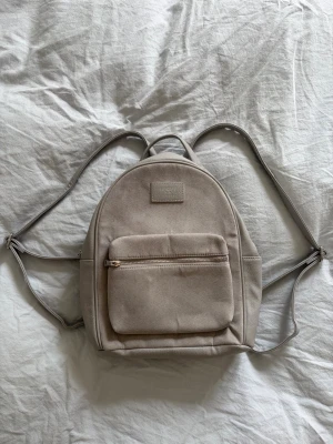 Beige/grå ryggsäck i syntetmaterial - Snygg lite mindre, beige/grå ryggsäck med enkel och stilren design. Väskan har ett stort huvudfack, en praktisk ytterficka med dragkedja och justerbara axelband. Tillverkad i ett skin/mocka likande material som ger en modern look. Perfekt för skola, stan eller utflykter.