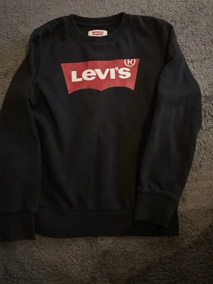 Svart sweatshirt från Levi's - Cool svart sweatshirt från Levi's med klassiskt rött och vitt logotryck på bröstet. Tröjan har rund halsringning och ribbade muddar vid ärmslut och nederkant. Perfekt för en avslappnad och trendig look.