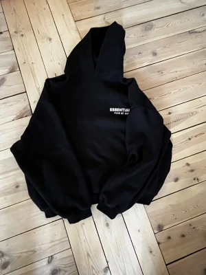 Svart Essentials Fear of God hoodie - Säljer en svart hoodie från Essentials Fear of God med vit logga på bröstet och stor text på ryggen. Hoodien har en klassisk huva och ribbade muddar vid ärmar och midja. Perfekt för dig som gillar streetwear och vill ha en clean look.