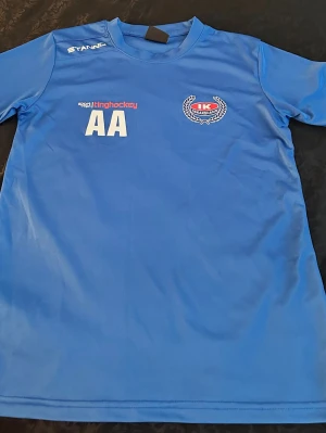Blå sporttröja från Stanno 152 cm  - Snygg blå sporttröja från Stanno med tryck på bröstet, inklusive klubbmärke och texten 'AA'. Tillverkad i ett lätt och funktionellt material som passar perfekt till träning eller match. Rund hals och korta ärmar för maximal rörelsefrihet.