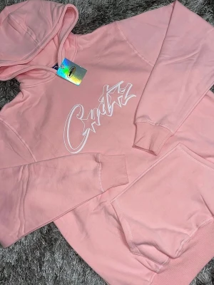 Corteiz allstarz  - Snygg rosa hoodie med vit text och stjärndetalj på bröstet. Hoodien har en stor magficka, ribbade muddar vid ärmar och nederkant samt en klassisk huva. Perfekt för dig som gillar streetwear och vill ha en chill look. Materialet känns mjukt och bekvämt.