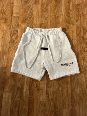 Grå Essentials shorts med snörning - Snygga ljusgrå shorts från Essentials med bred resår i midjan och långa snören framtill. Shortsen har två sidofickor och tryckt logga på vänster ben. Perfekta för chill dagar eller träning.