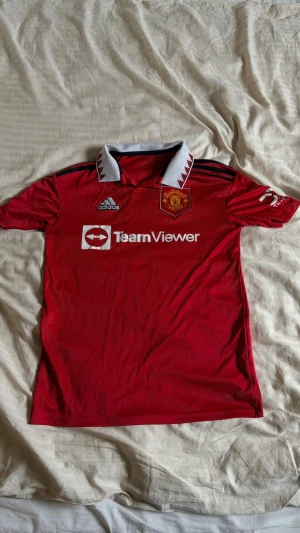 Manchester United matchtröja röd - Säljer en röd Manchester United matchtröja från Adidas med vit krage och röda detaljer. Tröjan har klubbmärke, Adidas-logga och TeamViewer-tryck på bröstet. Tillverkad i lätt och ventilerande material, perfekt för fotboll eller träning.