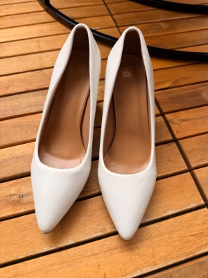 Vita klassiska pumps med spetsig tå - Säljer ett par stilrena vita pumps med spetsig tå och låg klack. Skorna har en slät yta i syntetmaterial och en neutral innersula i beige. Perfekta för dig som vill ha en clean och elegant look.
