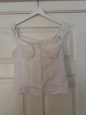 Vit blus med knappar och rynkad kant - Söt vit blus med korta ärmar och rynkad kant vid axlar och halsringning. Blusen har knappar framtill och en feminin passform som ger en romantisk vibe. Perfekt att styla med jeans eller kjol för en trendig look.