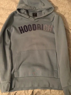 Ljusblå hoodie från Hoodrich XS - Snygg ljusblå hoodie från Hoodrich med broderad logga i lila och grått på bröstet. Klassisk känguruficka framtill och huva med metallringdetalj. Mjuk insida i fleece och relaxed fit. Perfekt för en chill och street-inspirerad look.
