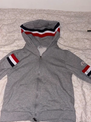 Grå Moncler hoodie med dragkedja - Snygg grå hoodie från Moncler med dragkedja framtill, huva med röd, vit och marinblå rand samt matchande detaljer på ärmarna. Klassisk Moncler-logga på ena ärmen. Perfekt för en chill och sportig stil.