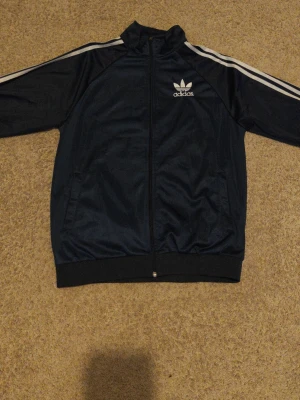 Marinblå Adidas vindjacka - Snygg marinblå vindjacka från Adidas med klassiska vita ränder längs ärmarna och Adidas-logga på bröstet. Jackan har dragkedja framtill, hög krage och ribbade muddar. Perfekt för en sportig och avslappnad stil.