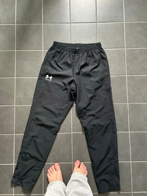 Svarta träningsbyxor Under Armour - Svarta träningsbyxor från Under Armour med resår i midjan och vit logga på vänster lår. Tillverkade i ett lätt, sportigt material med raka ben och enkel design. Perfekta för gymmet eller löprundan.