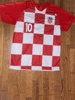 Kroatiens fotbollströja röd/vit - Säljer en fotbollströja med Kroatiens klassiska rödvita rutmönster, nummer 10 på bröstet och landslagets emblem. Tröjan har röda ärmar och V-ringning. Perfekt för dig som vill sticka ut på planen eller heja på laget.