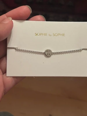 Silverarmband med peace-märke Sophie by Sophie  - Snyggt och stilrent armband i silver från SOPHIE by SOPHIE. Armbandet har en tunn kedja och ett runt hänge med peace-symbol. Perfekt för dig som gillar minimalistiska smycken med en cool detalj. Kan träffa i Göteborg 