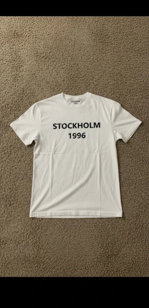 Vit S t-shirt STOCKHOLM 1996 - Eftertraktad Snygg vit t-shirt med svart tryck där det står STOCKHOLM 1996 på bröstet. Klassisk rund halsringning och korta ärmar. Tillverkad i mjuk bomull som känns skön mot huden. Perfekt för dig som gillar enkel och stilren design.