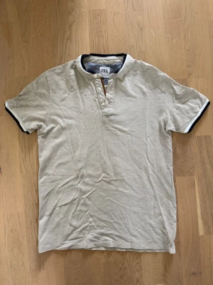 Beige kortärmad t-shirt från Zara - Snygg beige t-shirt från Zara med korta ärmar och kontrasterande mörkblå kant vid hals och ärmslut. T-shirten har knappar vid halsen för en extra detalj och är tillverkad i mjuk bomull. Perfekt för en avslappnad och stilren look.