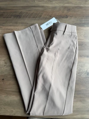 Beige kostymbyxor med pressveck - Snygga beige kostymbyxor med raka ben och pressveck. Byxorna har hällor för skärp, diskreta fickor bak och är tillverkade i ett mjukt, vävt material som ger en clean look. Perfekta för dig som gillar stilrena och klassiska plagg.
