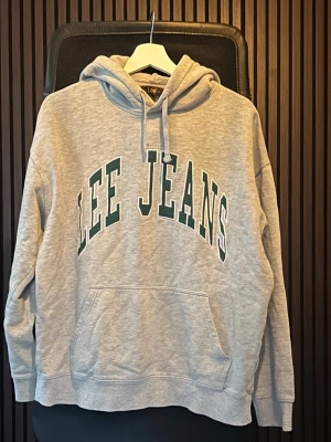 Grå hoodie från Lee Jeans i s - Snygg grå hoodie från Lee Jeans med stor logga i grönt och vitt på bröstet. Klassisk känguruficka framtill och justerbar huva med dragsko. Perfekt för en chill och avslappnad stil. Mjuk bomullsblandning och ribbade muddar vid ärmar och midja.