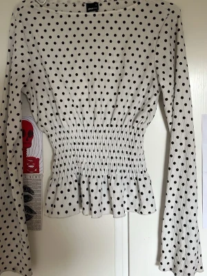 Vit prickig topp från Gina Tricot - Supersöt vit topp med svarta prickar från Gina Tricot. Toppen har långa ärmar, smockad midja och volang nedtill som ger en snygg siluett. Tillverkad i ett stretchigt och mjukt material som sitter bekvämt. Perfekt för dig som gillar trendiga detaljer.