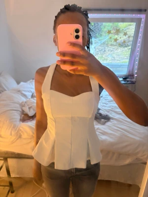 Vit korsettopp med halterneck - Säljer en snygg vit korsettopp med halterneck och figurnära passform. Toppen har en markerad midja och utsvängd nederkant som ger en cool siluett. Perfekt att styla med jeans eller kjol för en trendig look.