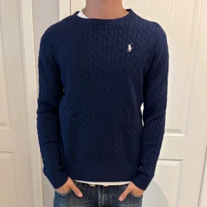 Mörkblå kabelstickad tröja Polo Ralph Lauren - Mörkblå kabelstickad tröja från Polo Ralph Lauren med rund hals och vit broderad logga på bröstet. Tröjan har långa ärmar, ribbade muddar och nederkant. Klassisk och tidlös design som passar till jeans eller chinos.