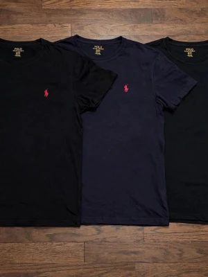Svarta och marinblå t-shirts Polo Ralph Lauren - Tre klassiska t-shirts från Polo Ralph Lauren i svart och marinblått med den ikoniska röda logon broderad på bröstet. T-shirtsen har rund hals och korta ärmar, perfekta för en clean och stilren look. Säljer billigt eftersom sommaren snart är över 