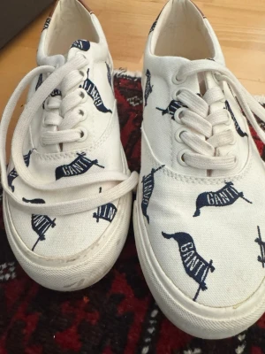 Vita sneakers med GANT-tryck - Snygga vita sneakers från GANT med marinblått flaggtryck över hela skon. Klassisk låg modell med vita snören och sula. Tillverkade i canvasmaterial som ger en avslappnad och trendig look. Perfekta för dig som gillar stilrena men ändå unika detaljer.