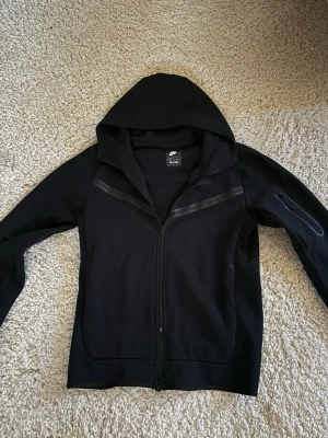 Svart Nike Tech Full-Zip Hoodie - Svart Nike Tech Full-Zip hoodie från Nike med hel dragkedja och huva. Har diskreta svarta detaljer över bröstet och Nike-logga på vänster sida. Tillverkad i mjukt material och har lång ärm. Perfekt för en sportig och avslappnad stil. Har bara andvänds två gånger. Alltså så gott som ny. 